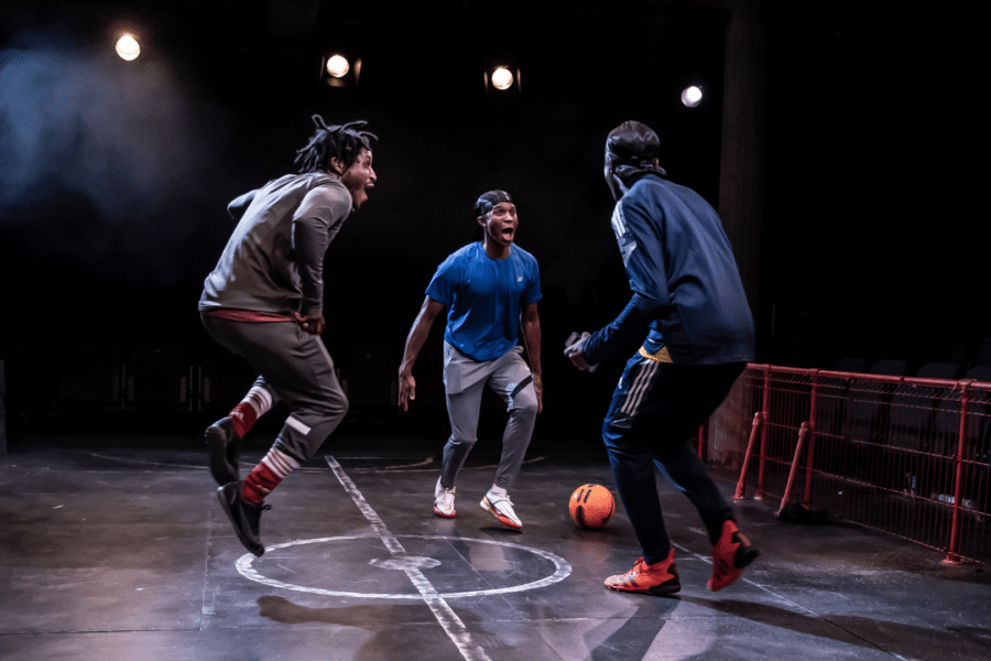 group of three people stood on stage facing each other with a football beside them, they look like they're all ready to jump in the air.