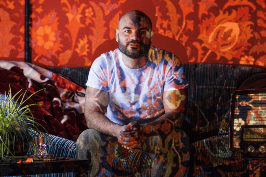 An Iraqi man sits on a sofa in a living room looking directly at a camera. There is a floral pattern being projected onto him and his surroundings.