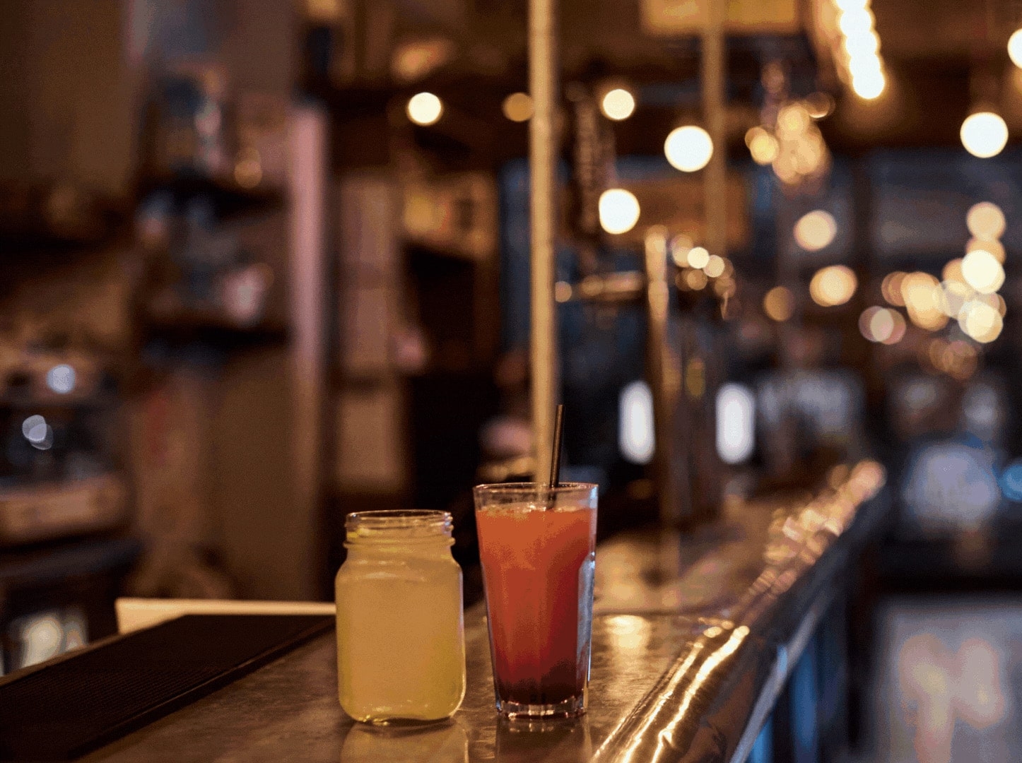 An image focusing on two cocktails on a bar. One is yellow and one is pink.