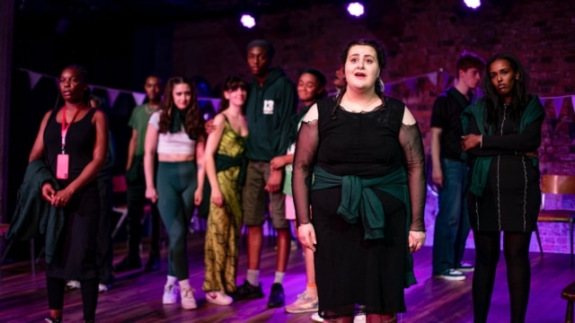 group of people on stage lit up purple, one person is stood closer to the front of the stage looking concerned.