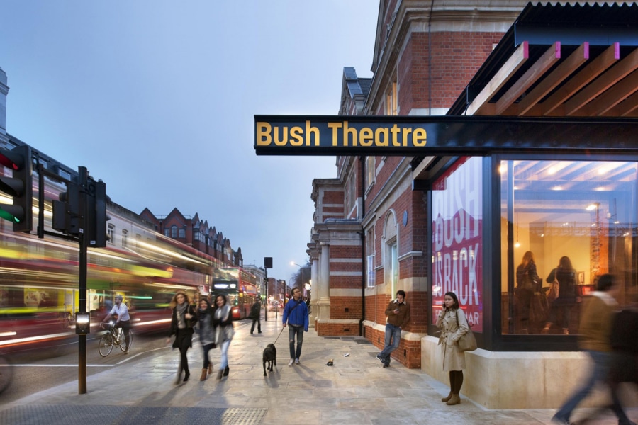 This is outside the Bush Theatre. There is a black sign with yellow writing stating Bush Theatre. There are many people outside walking around.