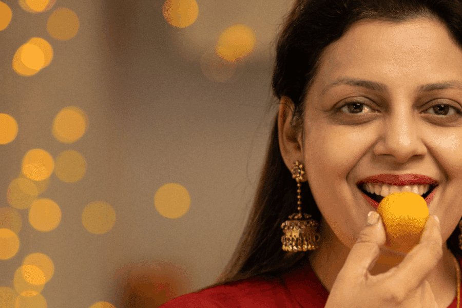 A South Asian woman wearing a red top and gold earrings holds her hand to her mouth whilst eating.