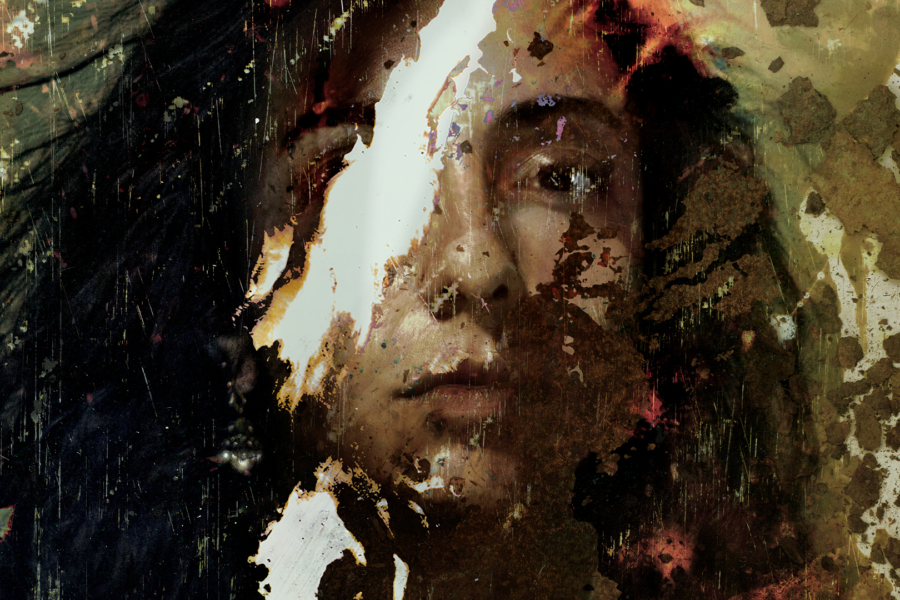 A south Asian woman with long black hair looks directly ahead. The photo is damaged with staining and scratches, with a large tear through her eye.