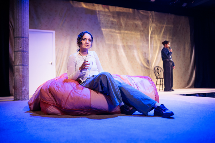 Production photo: Nadi Kemp-Sayfi sits on a mattress on the ground.