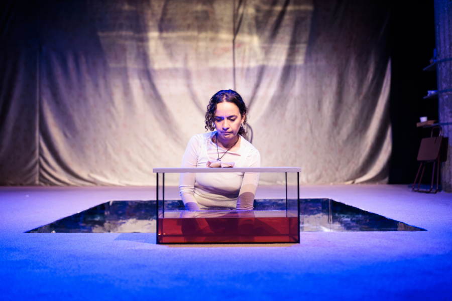 Production photo: Nadi Kemp-Sayfi sits on stage with a container of water in front of her. The water is coloured red with blood.