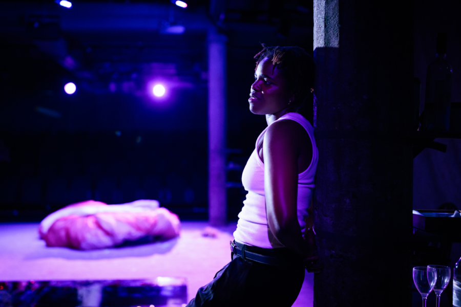 Production photo: Francesca Amewudah-Rivers stands with her back to a pillar, lit by purple light.
