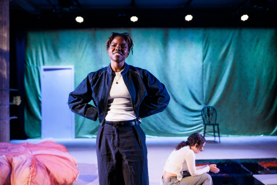 Production photo: Francesca Amewudah-Rivers stands with her hands on her hips. She is smiling and Nadi Kemp-Sayfi sits in the background.