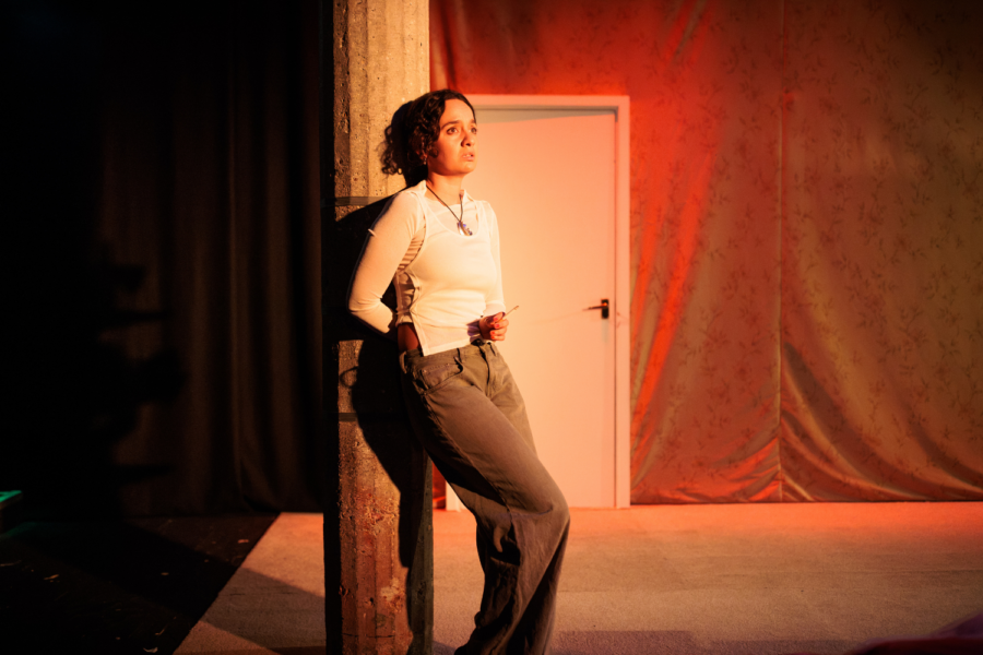 Production photo: Nadi Kemp-Sayfi leans back against a pillar, illuminated by warm red light.