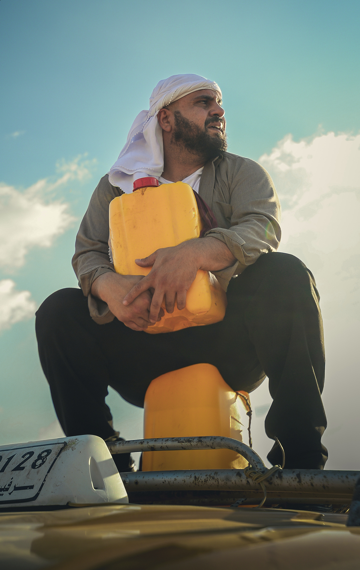 Alaa Shehada sits on a yellow container atop a bus. He holds another yellow container and looks off to the side.